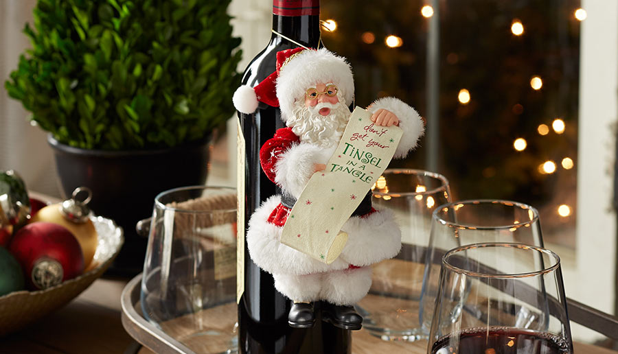 Decorative Santa Claus figure on a wine bottle with glasses and Christmas decorations in the background.